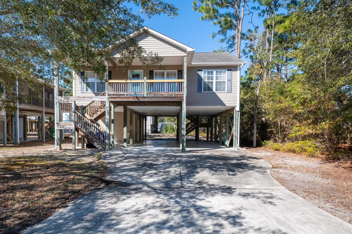 This image showcases a charming elevated single-family house nestled among mature trees, with a spacious front porch overlooking a private driveway. The welcoming scene is set for relaxing mornings with coffee in hand or enjoying sunset views with loved ones. The peaceful, wooded setting hints at both seclusion and easy access to Oak Island's beach.