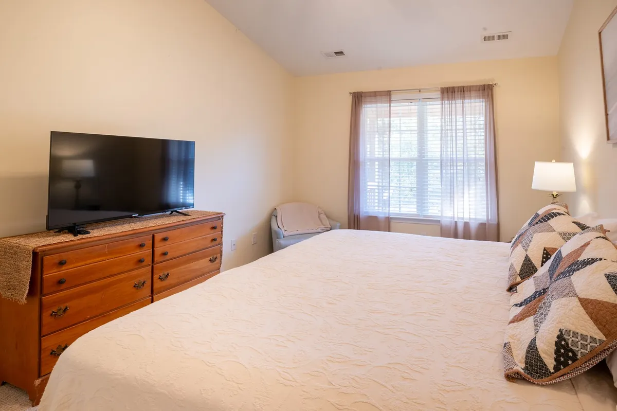 A spacious bedroom featuring a comfortable bed with neutral linens, a dresser, a smart TV, and a window with curtains that allow natural light to filter in. The serene, tranquil mood and functional elements like the TV and dresser make it ideal for relaxing, watching TV, or unwinding after a long day. This room likely represents the primary suite described in the section.