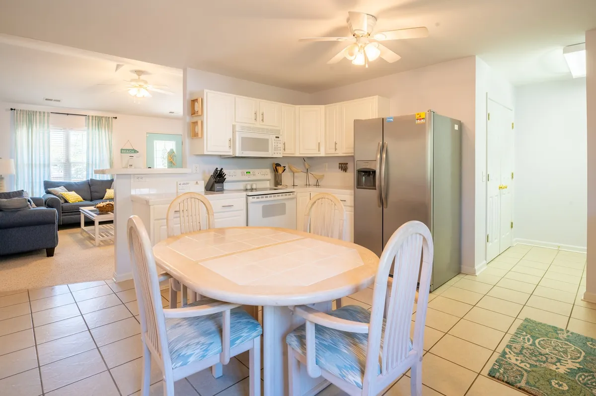 The view from the dining area into the kitchen, with a dining table set for a family meal and kitchen appliances in the background. The open floor plan is evident, seamlessly connecting the dining, kitchen, and living zones. The warm lighting and layout invite guests to share meals and conversations together.