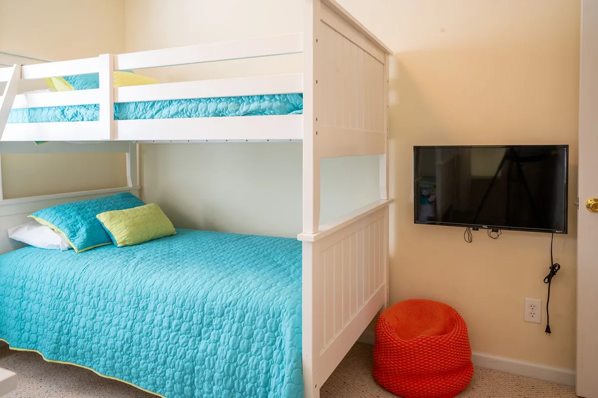 A playful kids' room with twin bunk beds dressed in colorful bedding, a TV, a cheerful orange pouf, and a window letting in natural light. The lively atmosphere makes it ideal for sleep, play, and movie nights, and the safe, child-friendly design is perfect for families with little ones.
