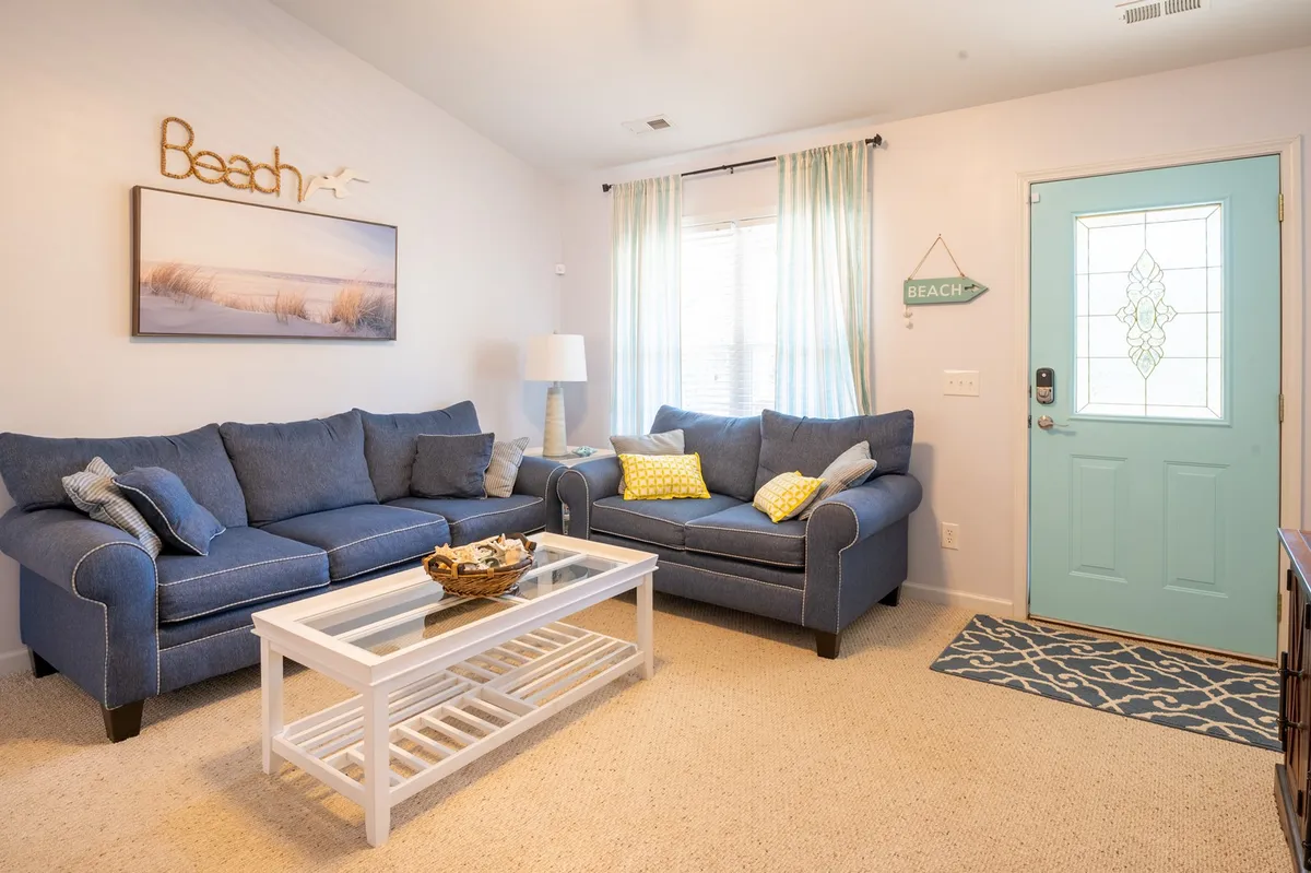 A cozy, open-concept living area featuring plush blue sofas, a white coffee table, and beach-themed wall art. The turquoise door adds a pop of color, while natural light streams in to create a welcoming, cheerful atmosphere. The space is clearly designed for relaxation and socializing, with ample room for guests to gather.