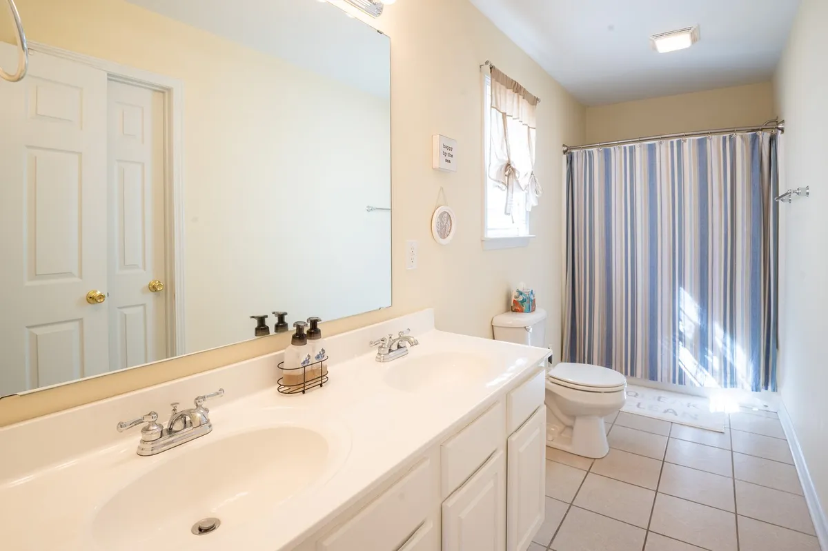 A clean, modern bathroom with double sinks, a large mirror, a striped shower curtain, and a toilet. The calm and organized mood makes it perfect for morning routines or relaxing baths, and the spacious layout supports family convenience.