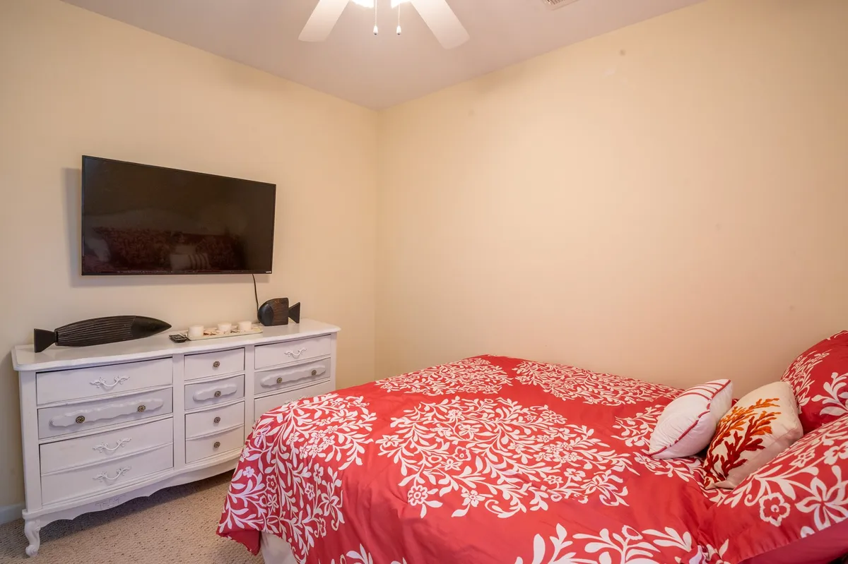 A bright and inviting bedroom with a bold red floral bedspread, a white dresser, and a flat-screen TV. The cozy and welcoming mood is perfect for restful sleep and quiet evenings. This room is well-suited to represent the queen bedroom described in the section.