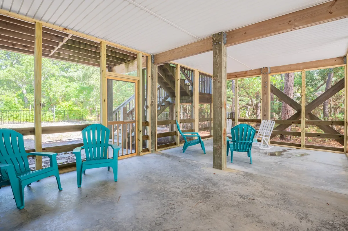 A bright and welcoming covered porch with turquoise Adirondack chairs set beneath sturdy wooden support beams. Sunlight filters through nearby live oaks, casting soft shadows. The porch feels open and airy, perfect for enjoying a peaceful morning coffee or relaxing with friends and family. A staircase leads up, suggesting a connection to the rest of the home.